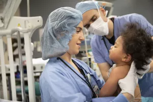 Nurse holding pediatric baby patient in operating room and smiling while team member assists in background.